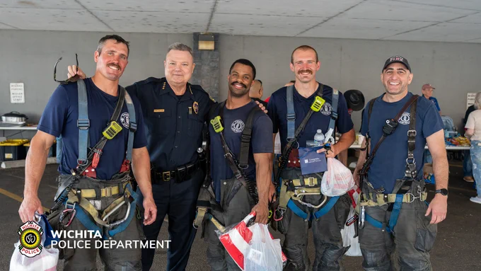 A group of five individuals, including four in matching rescue harnesses and one in a police uniform, stand together smiling. They appear to be in an outdoor area with a concrete ceiling. Visible logos identify the Wichita Police Department. The individuals are holding various items like bags and drinks. Other people and tables are partially visible in the background.