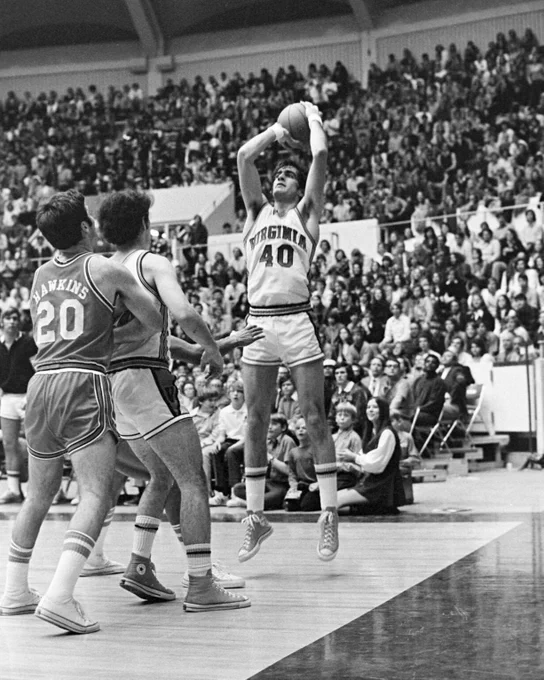A UVA basketball player wearing a "Virginia" jersey with the number 40 is seen taking a jump shot during a game. Two opposing players are near, one wearing a jersey with the number 20. The scene is set in University Hall with spectators filling the stands.