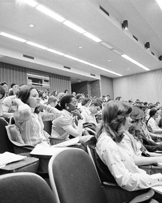 A packed lecture hall at UVA filled with students attentively listening and taking notes. The room has tiered seating with several rows visible, and students appear engaged with the lecture. The interior has brick and panel walls with overhead lighting.