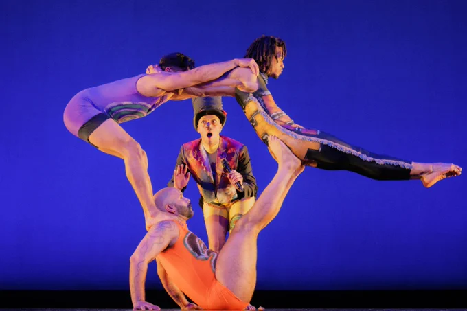 Three Minty Fresh Circus performers are balancing in a striking acrobatic pose on stage against a blue background. Another performer stands nearby, dressed in a colorful costume with a top hat.