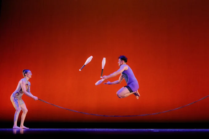 Two Minty Fresh Circus performers are on stage against a bright red backdrop. One is spinning a jump rope while the other is skillfully juggling clubs and jumping over the rope.