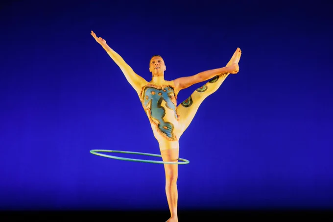 A Minty Fresh Circus performer balances on one foot, extending the other leg high, while spinning a glowing hula hoop. The stage backdrop is a deep blue. The performer wears a vibrant bodysuit with an intricate design.