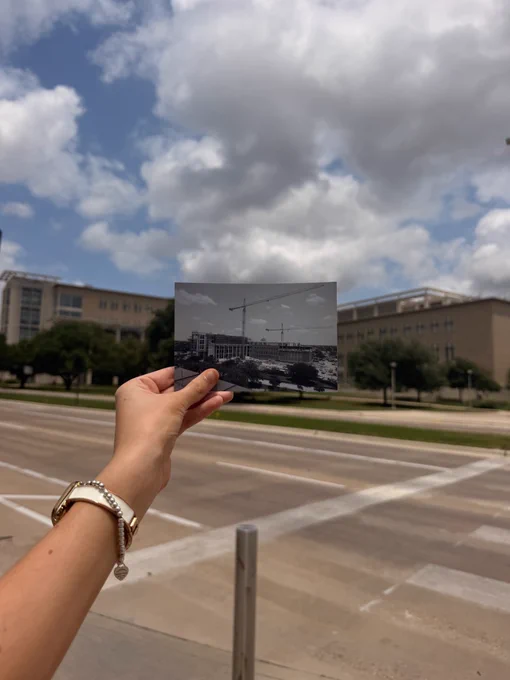 A hand holds a black and white photo in front of a modern street scene. The photo depicts a construction site with cranes, similar to the current background. Buildings and cloudy skies appear in both the image and the background.