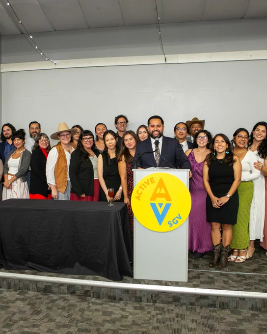 A group of people standing together, some smiling, around a podium with the "Active SGV" logo on it. The individuals are dressed in formal and semi-formal attire. The backdrop is a plain gray wall and there is a long table covered with a black cloth next to the group.