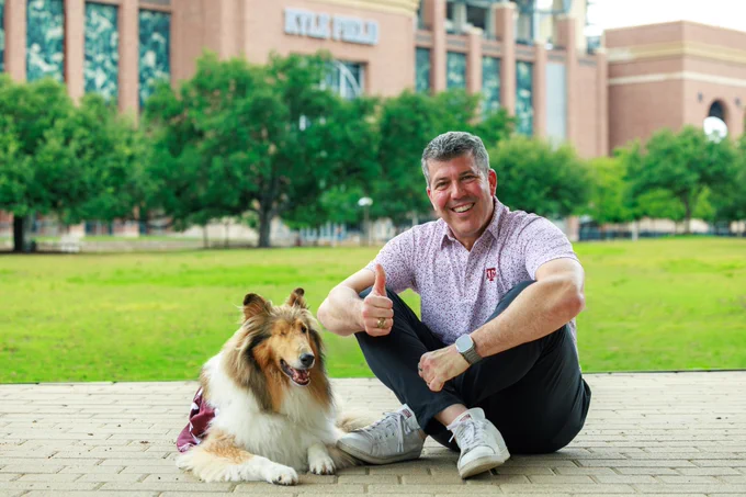 A person sits on a walkway, giving a thumbs up, with a collie wearing a maroon blanket beside them. In the background is Kyle Field surrounded by trees.