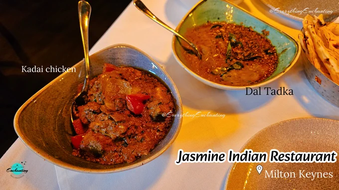 An angled, top-down photograph of an Indian dinner spread arranged on a white tablecloth at Jasmine Indian Restaurant in Milton Keynes. In the foreground on the left, a triangular grey-rimmed bowl is filled with Kadai Chicken, featuring tender meat cooked in a thick, rich gravy with visible pieces of bell pepper and a serving spoon resting inside. To the upper right, a similar light-green triangular bowl holds Dal Tadka garnished with whole spices and fresh herbs. Part of a bread basket with naan or roti is visible on the far right. The image contains white text labels reading "Kadai chicken" and "Dal Tadka", along with a stylized footer that says "Jasmine Indian Restaurant" next to a location pin for "Milton Keynes". The watermark "@EverythingEnchanting" and a circular blog logo are also present.
