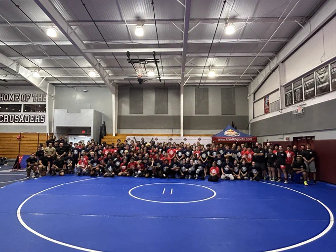 A large group of people gathered in a gymnasium with a wrestling mat and banners. The mood appears energetic and communal.