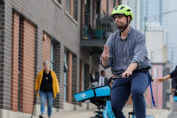 Riley O'Neil riding a Divvy bike, wearing a helmet, giving a "peace" sign with right hand