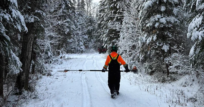 A staff member walking down a snowy trail through the forest, wearing high visibility shirt, winter clothes, and PPE, while carrying a trail maintenance tool. 