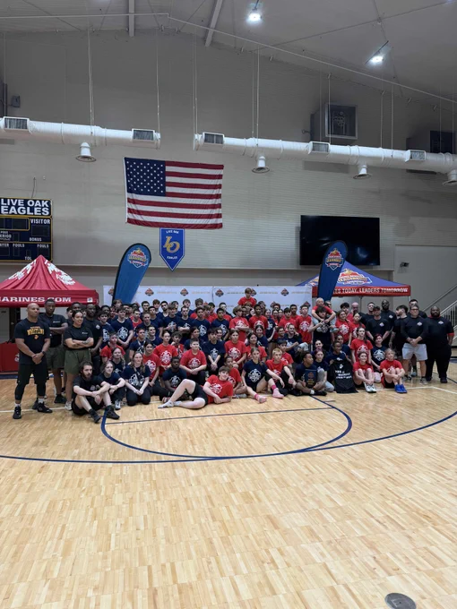 A large group of people are gathered in a gymnasium beneath an American flag. They appear to be part of an organized event and are wearing matching red and black shirts.