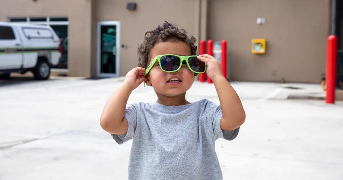 A young child stands outdoors on a paved surface, adjusting large, bright green sunglasses. In the background, there is a beige building with visible doors, a white vehicle, and red bollards.