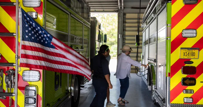A person in a hat and another individual are in a fire station between two bright yellow and red fire trucks. One fire truck displays an American flag. Light filters through the open entrance in the background.