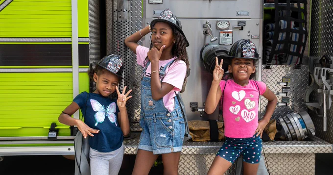 Three children pose in front of a bright yellow fire truck, wearing firefighter helmets. The child on the left is making a peace sign, the middle child is striking a thoughtful pose, and the child on the right has fingers in a peace sign.