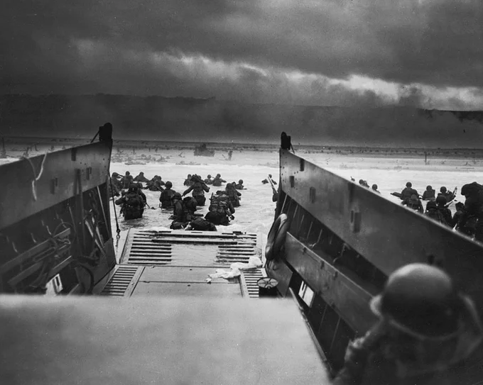 A historic World War II scene depicts soldiers disembarking from a landing craft onto Omaha Beach during D-Day. The sky is overcast, and smoke is visible in the background, suggesting the intensity of the battle.