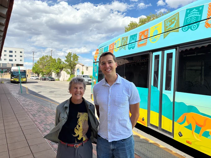 Two TAB members standing on a sidewalk next to a colorful bus with animal and geometric designs under a partly cloudy sky.