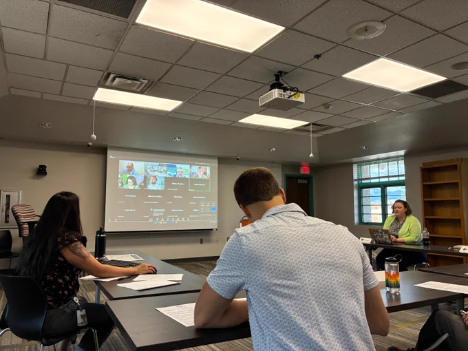 Three people in a conference room attending a hybrid meeting with a video call projected on a screen.