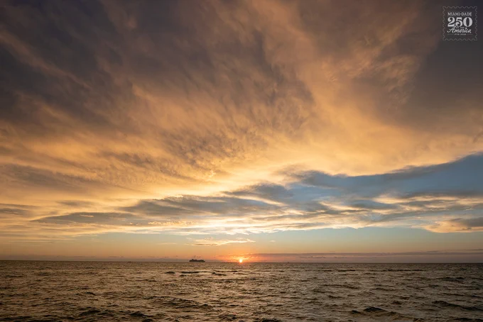 Sunset over a vast ocean with dramatic golden and orange clouds streaked across the sky. The sun is setting on the horizon, casting a bright glow, with a large ship silhouetted in the distance. The water is dark and wavy. Top right: \"MIAMI-DADE 250 America 1776-2026.\"