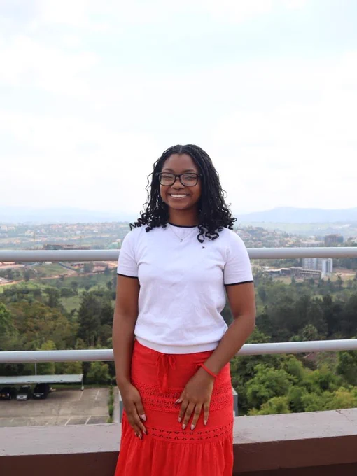 A person stands on a balcony with a scenic view of a lush, green landscape and distant mountains. They are wearing glasses, a white shirt, and a red skirt, smiling at the camera.