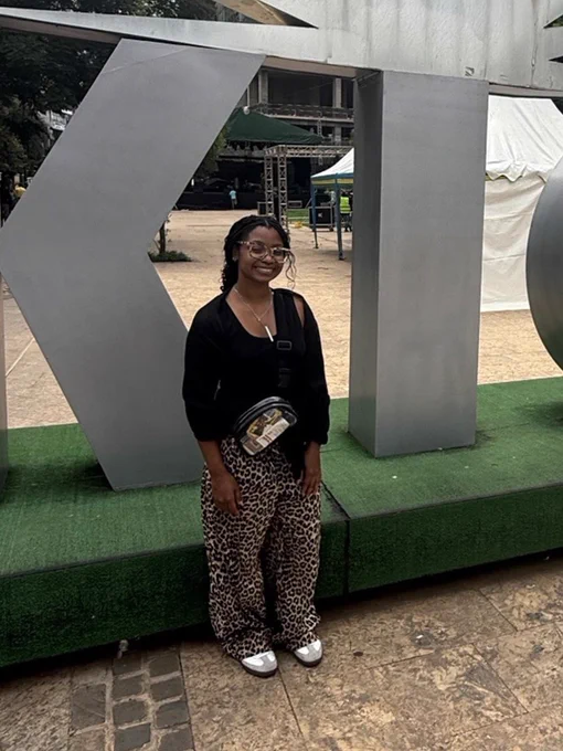 A person standing in front of large metallic letters "KIG" in an outdoor setting. The individual is wearing glasses, a black top, leopard-print pants, and carries a black crossbody bag. Trees and a building are visible in the background.