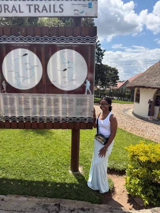 A person stands smiling beside a large informational sign about "Royal Trails" and "Big View Trail." The setting is outdoors, with green grass and a thatched-roof building in the background under a partly cloudy sky.
