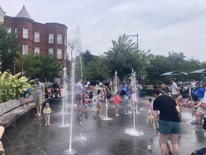 Children and adults enjoying a splash pad with multiple water jets shooting up from the ground in a lively urban park setting, surrounded by benches, greenery, and historic brick buildings under a cloudy sky.