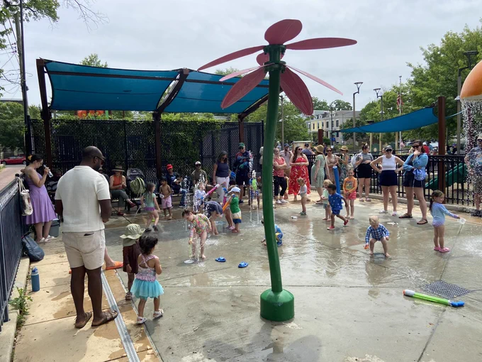 Children and adults enjoying a splash pad water play area on a cloudy day, with kids running through water jets and a large flower-shaped water feature in the center, while parents watch nearby under blue shade canopies.