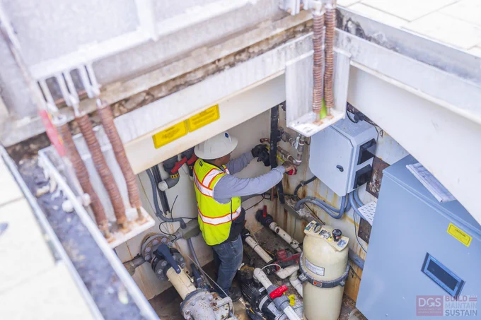 Technician wearing a safety helmet and high-visibility vest working inside a confined utility pit surrounded by pipes, valves, and control panels, performing maintenance or inspection tasks.