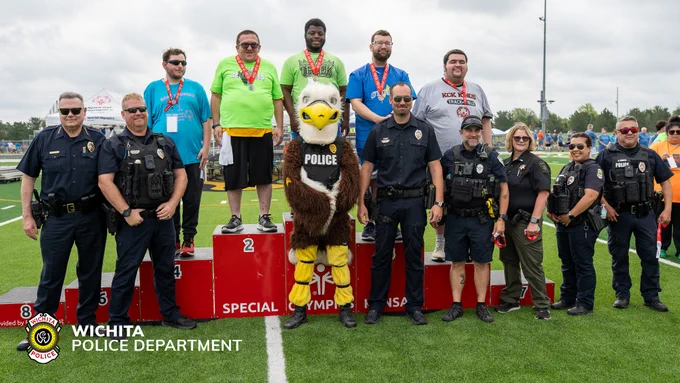 The image shows a group of individuals, including participants wearing medals, standing on podiums numbered 2 and 3. A person in a bird costume labeled "Police" stands in the center. Several police officers are positioned around them. The scene takes place outdoors on a grassy field. A Wichita Police Department logo is visible in the bottom left corner.