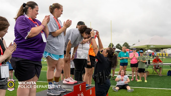 The image shows a person in a Wichita Police Department uniform awarding medals to athletes standing on a podium at a sports field. Other individuals are applauding, and a field with goalposts is visible in the background. The scene appears to be celebratory.