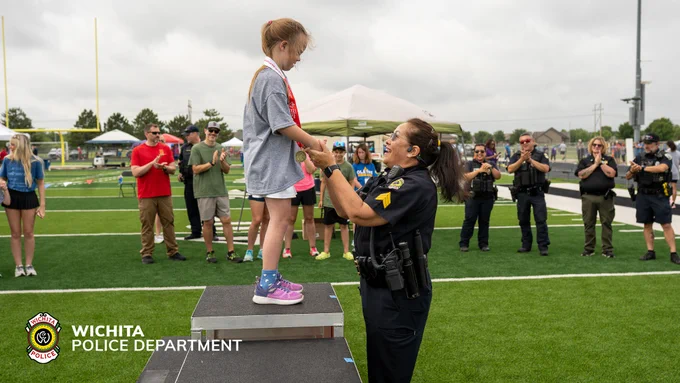 A young person stands on a podium receiving a medal from a Wichita Police Department officer at an outdoor event on a sports field. Several people in the background, including other officers and attendees, are applauding. The Wichita Police Department logo is visible.
