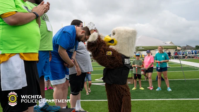 A person in blue sports attire is being awarded a medal by a mascot dressed as an eagle from the Wichita Police Department on a sports field. Other individuals are standing nearby, some clapping or observing. There is a tent in the background.