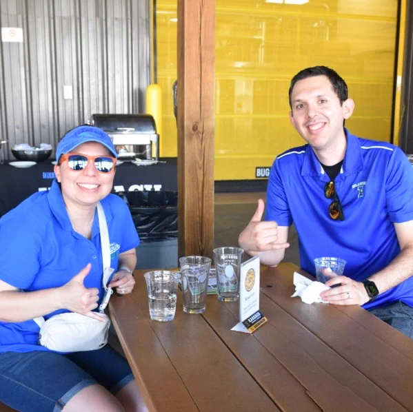 Two Texas A&M University-Corpus Christi Islander alumni seated at a wooden table, both wearing blue TAMU-CC Islanders shirts. They are making a friendly "shakas up" hand gesture. Several empty glasses and a menu are on the table. The background features a metallic wall and a bright yellow section.