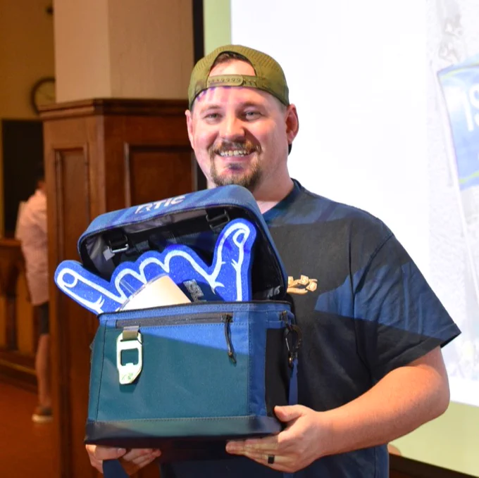 A Texas A&M University-Corpus Christi alumnus smiling while holding a blue YETI cooler containing a foam hand making a "shakas up" gesture and a can opener attached to the cooler. The person is wearing a cap backwards.