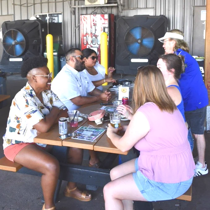 A group of Texas A&M University-Corpus Christi alumni sit around a picnic table, enjoying drinks and food. They are in an outdoor setting with large fans behind them. Some food containers and drinks can be seen on the table.