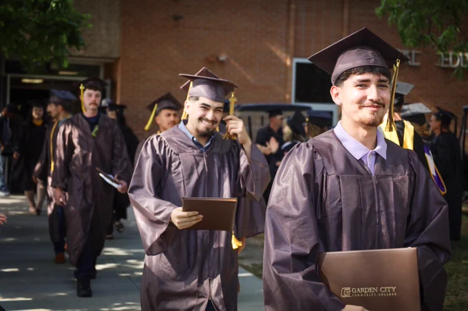 Graduates in caps and gowns holding diplomas while walking outside a brick building during a commencement ceremony.