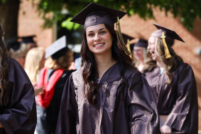 Graduate wearing a black cap and gown standing outdoors with blurred graduates and guests in the background.
