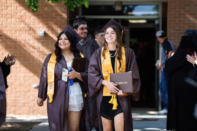 Group of graduates wearing caps and gowns walking outside a brick building during a commencement ceremony.