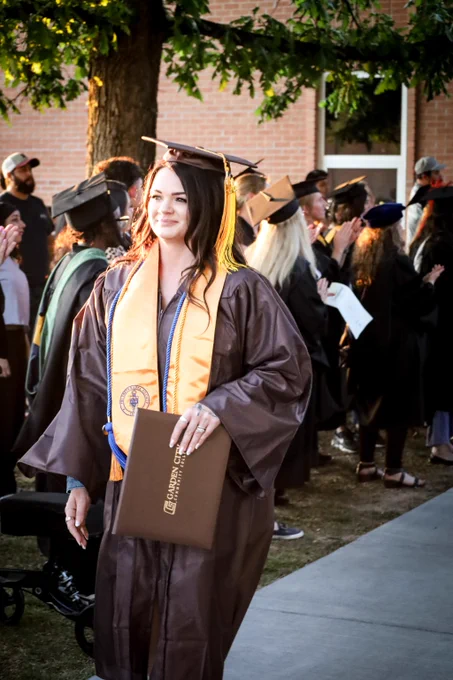 Graduate in brown cap and gown holding diploma cover during outdoor commencement ceremony with peers in background.