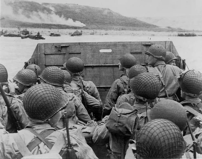 American assault troops in a landing craft huddle behind the protective front of the craft as it nears a beachhead on the northern coast of France on June 6, 1944. Smoke in the background is naval gunfire supporting the landing.