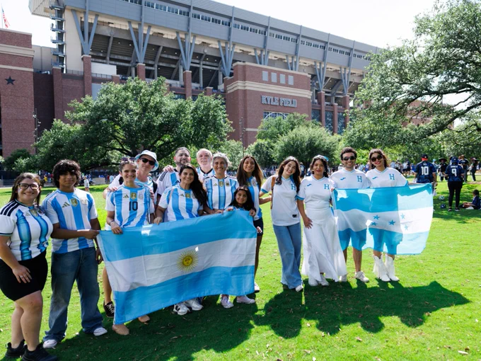 A group of people stand together on a grassy area, holding the flags of Argentina and Honduras. They are in front of Kyle Field, with a large building and trees visible in the background. The group appears to be celebrating or attending an event.