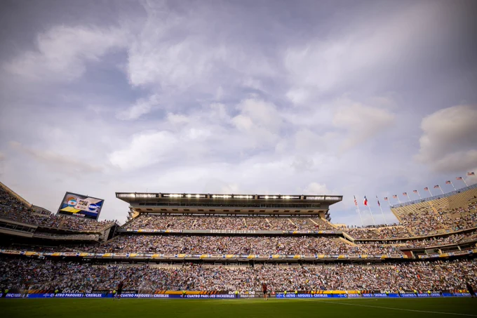 A large crowd fills Kyle Field, with a prominent scoreboard and flags visible under a partly cloudy sky.