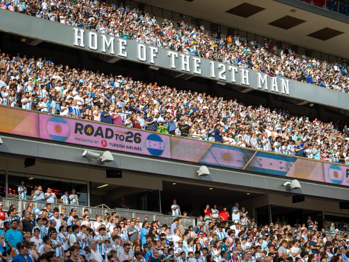 A large, enthusiastic crowd fills the seats of a stadium. A prominent banner reads, "Home of the 12th Man." Another banner below, displaying the logo of Turkish Airlines, says, "Road to 26." The atmosphere is lively and packed with people.