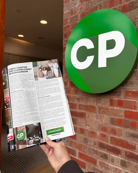Hand holding the magazine article beside a green CP logo sign mounted on a brick wall.