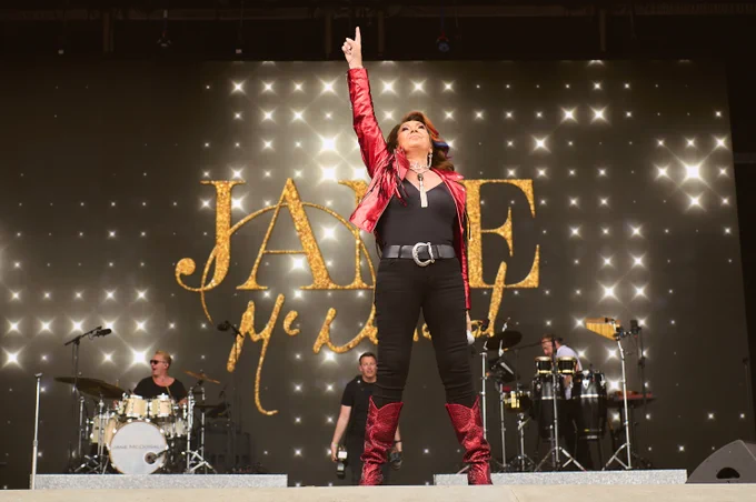 A performer stands confidently on stage with one arm raised, wearing a red outfit and boots. In the background, a band is playing, and a large screen displays the name "JANE McDONALD" in bold, gold letters.