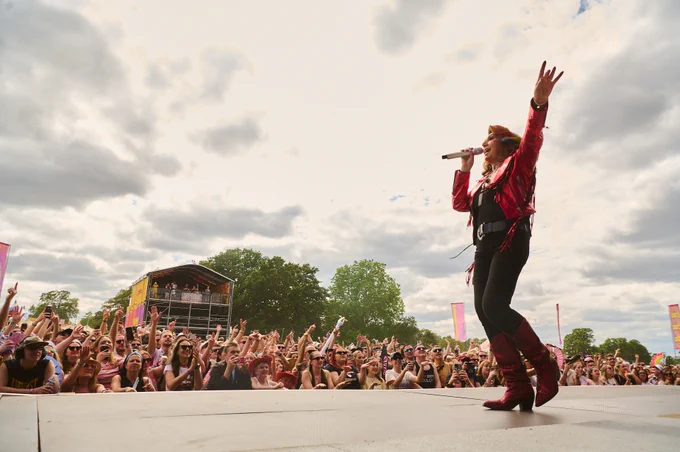 A performer on stage at a music festival, engaging a large, enthusiastic crowd. The audience is cheering with arms raised, and a structure in the background holds additional spectators. The sky is cloudy with some patches of blue.