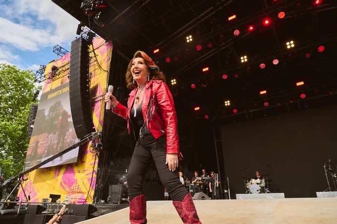 A performer in a red jacket and cowboy boots sings energetically on an outdoor stage. A large screen displays them, set against vivid pink and yellow graphics. Musicians can be seen in the background, with trees and a bright sky visible on the left.
