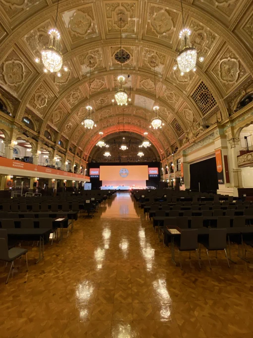 Image of a ballroom dance hall, with GMB branding around it and seats lined up either side of an aisle.