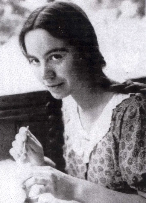 Historical photograph of a woman smiling subtly at the camera while sitting outdoors on a bench, holding an object in her hands.
