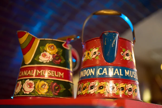 Two vintage painted metal water cans with floral rosette motifs, bright red and green paint and "Canal Museum" lettering.