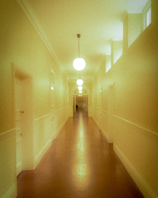 A long, warmly lit hallway with polished wooden floors and round ceiling lights. The corridor is lined with white doors and has high windows near the ceiling.